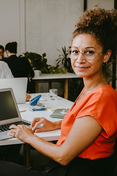 Eine Frau in roter Bluse lächelt freundlich in die Kamera, während sie an einem Schreibtisch arbeitet.