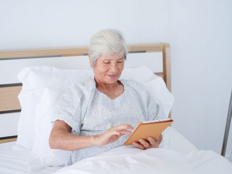 Eine ältere Dame bedient ein Tablet in einem Pflegebett.